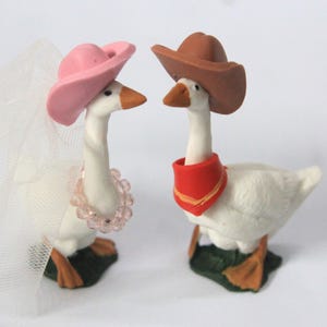 May include: Two white goose figurines standing on green bases. The left goose wears a pink cowboy hat, a beaded necklace, and a white tulle veil. The right goose wears a brown cowboy hat and a red bandana around its neck. Both have orange beaks.