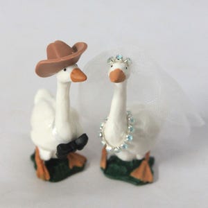 May include: Two white goose figurines standing on green bases. The left goose wears a brown cowboy hat and a black bow tie. The right goose wears a white tulle veil with a small crown of blue beads and a matching beaded necklace around its neck.