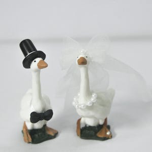 May include: Two white goose figurines, one wearing a black top hat and bow tie, the other adorned with a pearl necklace and a white veil. The geese are standing on a green base, perfect for a wedding cake topper.