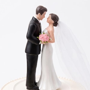 Puede incluir: Un adorno para pastel de bodas con una figura de novia y novio. El novio lleva un esmoquin negro, y la novia un vestido blanco, sosteniendo un ramo rosa. Opciones de color de cabello: rubio, castaño y negro. El adorno está sobre una base de pastel blanca.