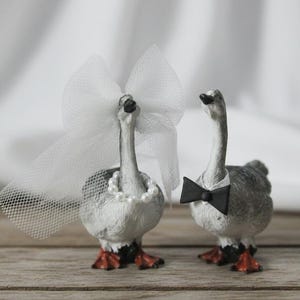 May include: Two goose figurines, one adorned with a white veil and pearl necklace, the other with a black bow tie. The geese have gray and white feathers, orange feet, and are likely wedding cake toppers.