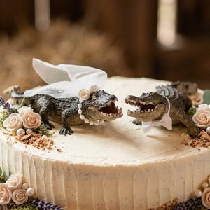 May include: A round cake topped with two alligator figurines. One alligator wears a white tulle veil and a pearl necklace, while the other wears a white bow tie. The cake is decorated with light pink roses, small white pearls, green leaves, and lavender sprigs.