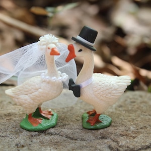 May include: A pair of whimsical goose figurines, one adorned with a bridal veil and pearl necklace, the other with a top hat and bow tie. The geese stand on green bases, perfect for a wedding cake topper.