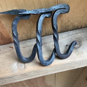 May include: Hand-forged metal wall hook with a unique design. The hook features three curved, twisted hooks and a flat top piece. The metal has a dark, rustic finish, perfect for a farmhouse or industrial decor style.
