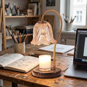 May include: A decorative table lamp with a flower-shaped shade in soft pink. The lamp has a gold-colored curved stem and sits on a dark, round base. A lit candle in a glass holder is on the base. A laptop and open sketchbook are nearby.