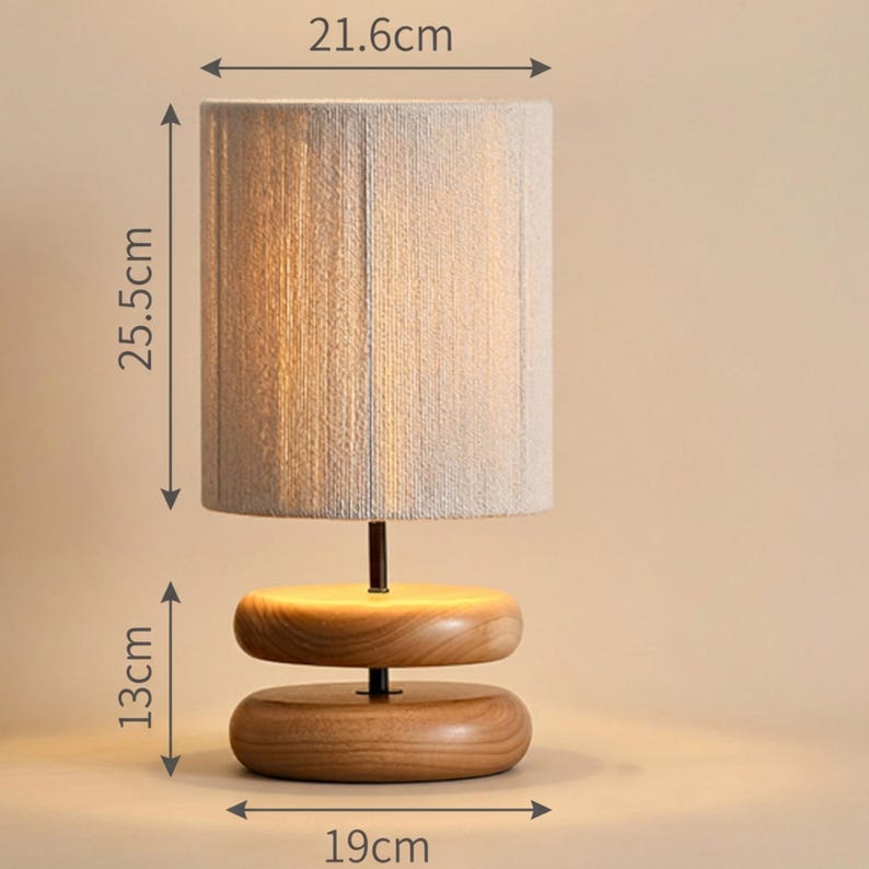 May include: A modern table lamp with a cylindrical, light-colored fabric shade, 21.6cm wide and 25.5cm tall, atop a wooden base. The base features two stacked, rounded wooden pieces, 19cm wide and 13cm tall, connected by a dark metal rod.