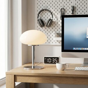 May include: A modern desk setup featuring a white mushroom-shaped table lamp with a chrome base, a digital clock displaying 10:35 AM, and a white coffee cup. A computer monitor, headphones, and a pegboard are in the background.