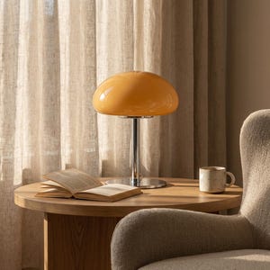 May include: A retro-style table lamp with a chrome base and a rounded, amber-colored glass shade. The lamp sits on a wooden side table next to an open book and a ceramic mug. The background features a neutral-toned curtain and a cozy armchair.