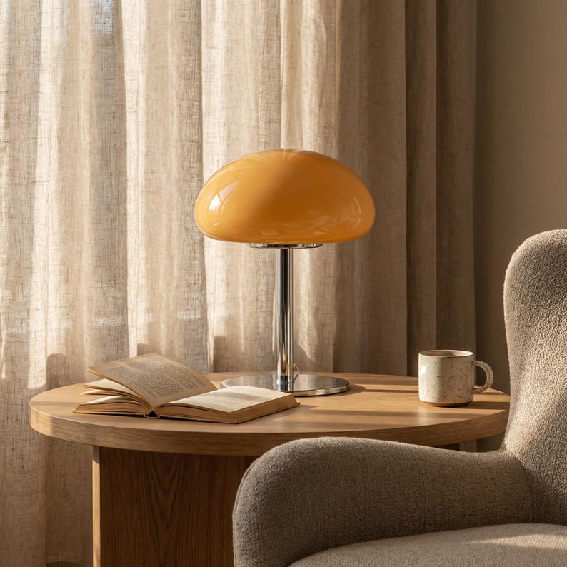 May include: A retro-style table lamp with a chrome base and a rounded, amber-colored glass shade. The lamp sits on a wooden side table next to an open book and a ceramic mug. The background features a neutral-toned curtain and a cozy armchair.