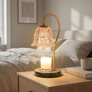 May include: A decorative table lamp with a flower-shaped glass shade and a gold-colored metal frame. The lamp sits on a green marble base, with a lit candle inside. The lamp is on a wooden nightstand next to a bed.