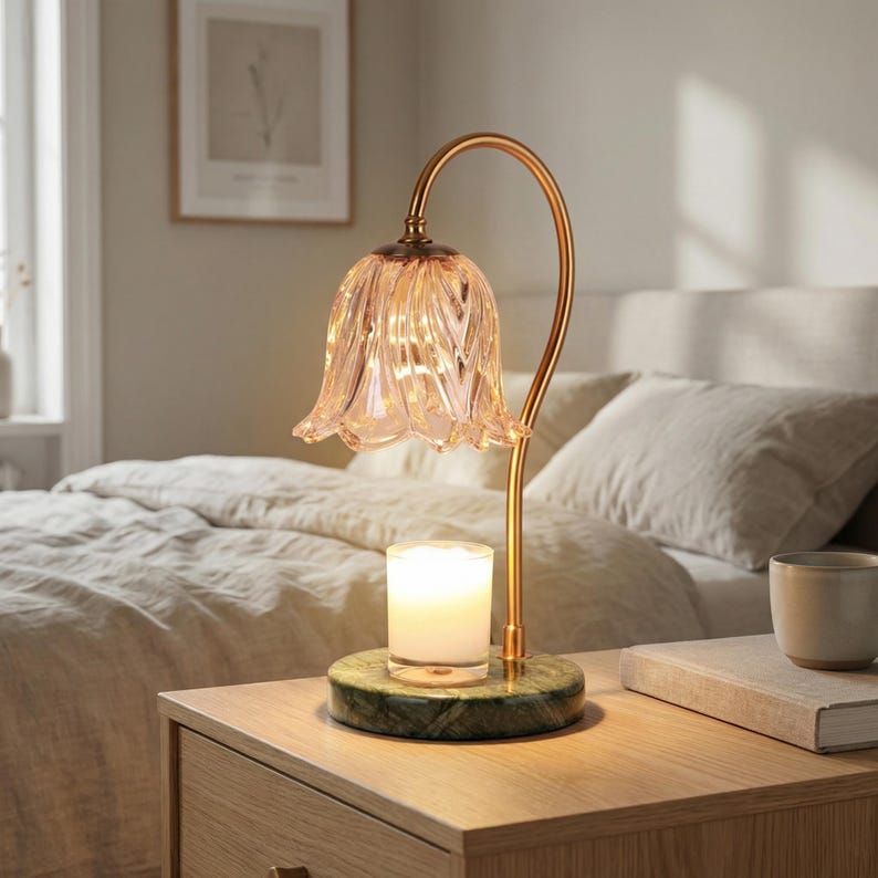 May include: A decorative table lamp with a flower-shaped glass shade and a gold-colored metal frame. The lamp sits on a green marble base, with a lit candle inside. The lamp is on a wooden nightstand next to a bed.