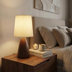 Puede incluir: Una mesita de noche de madera con una lámpara, libros, una taza y una vela. La lámpara tiene una pantalla plisada y una base de madera oscura. La taza y la vela son de color beige. Los libros están apilados.