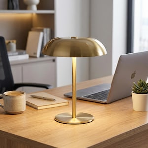 Puede incluir: Una l&aacute;mpara de escritorio dorada con una pantalla redondeada y un tallo delgado. La l&aacute;mpara est&aacute; sobre un escritorio de madera, junto a un port&aacute;til, una taza de caf&eacute; y una peque&ntilde;a planta en maceta. El dise&ntilde;o de la l&aacute;mpara es minimalista y moderno.