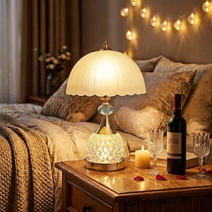 Peut inclure: Une lampe de table décorative avec un abat-jour festonné crème. La lampe a une base en cristal et une tige dorée. Une bouteille de vin rouge, deux verres à vin et une bougie allumée sont posés sur une table de chevet en bois.