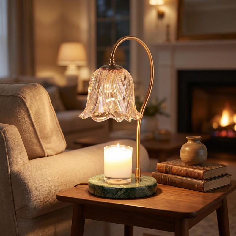May include: A decorative table lamp with a clear, flower-shaped glass shade and a gold-colored curved stem. The lamp sits on a green marble base, with a lit candle. A small wooden table holds the lamp, books, and a vase.