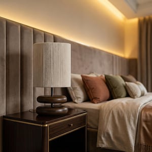 May include: A bedroom scene featuring a dark wood nightstand with a lamp and a bed with pillows. The lamp has a beige fabric shade and a dark wood base. The bed has a beige comforter and several pillows in shades of beige, orange, and green.