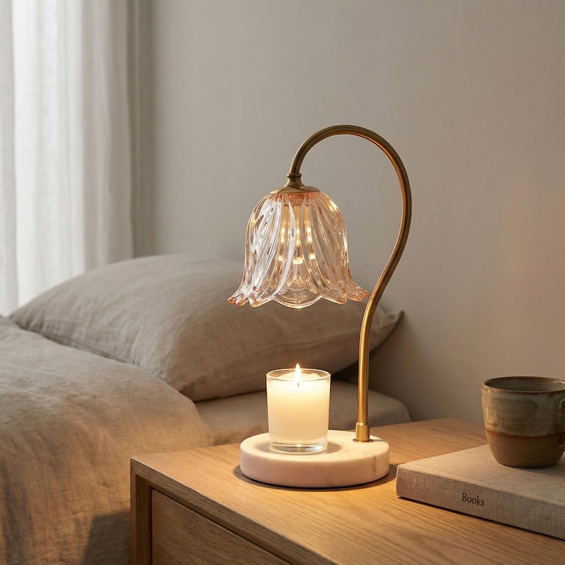 May include: A decorative bedside lamp with a flower-shaped, pink-tinted glass shade and a curved brass stem. A lit candle sits on a marble base below the lamp. A ceramic mug and a book are on the wooden nightstand.