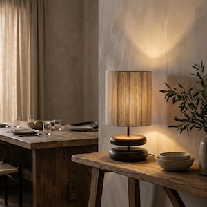 May include: A wooden table lamp with a cylindrical lampshade and a dark brown base. The lampshade is a light brown color. The lamp is on a rustic wooden table. The background includes a dining table and a vase with greenery.