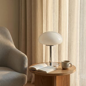 May include: A white mushroom-shaped table lamp with a chrome base sits on a round wooden side table. An open book and a mug of coffee are also on the table. A gray armchair is to the left, and beige curtains are in the background.