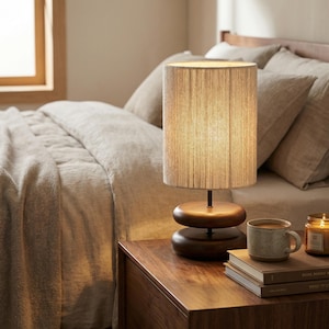 May include: A wooden bedside table with a lamp, books, and a mug. The lamp has a cylindrical shade and a dark brown base. A lit candle sits next to a mug of coffee and a stack of books. The bedding is a neutral color.