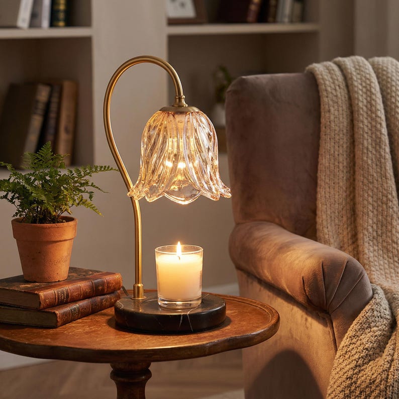 May include: A decorative table lamp with a gold-colored curved stem and a clear, flower-shaped glass shade. A lit candle sits on a black marble base. The lamp is on a wooden table with books and a potted plant.
