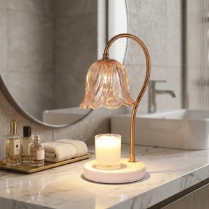 May include: A decorative candle holder with a pink glass flower-shaped shade and a gold-colored metal stem. The candle holder sits on a white marble base. The candle holder is on a marble countertop in a bathroom setting.