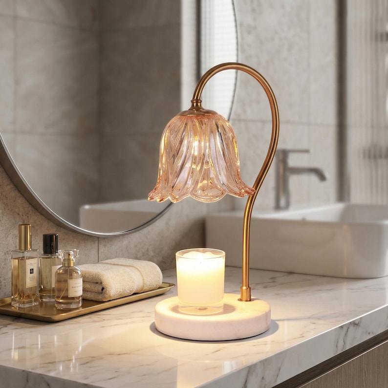 May include: A decorative candle holder with a pink glass flower-shaped shade and a gold-colored metal stem. The candle holder sits on a white marble base. The candle holder is on a marble countertop in a bathroom setting.