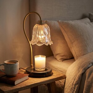 May include: A decorative bedside lamp with a clear, flower-shaped glass shade and a gold-colored metal stem. A lit candle sits on a black marble base. A mug and a book are on a wooden nightstand.