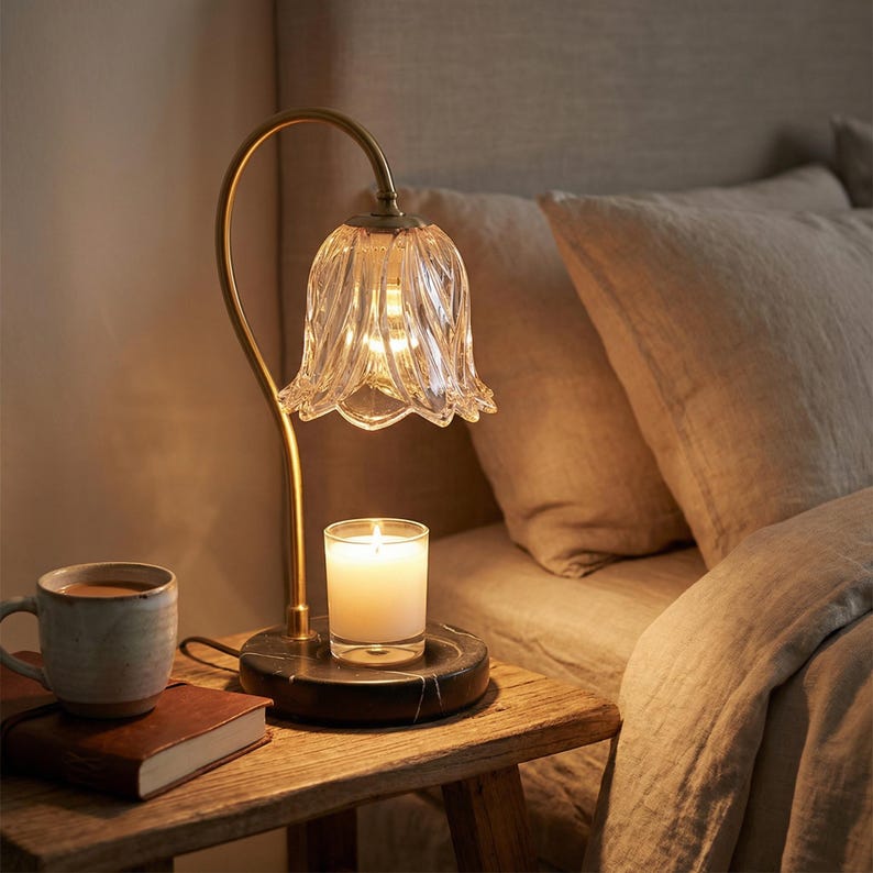 May include: A decorative bedside lamp with a clear, flower-shaped glass shade and a gold-colored metal stem. A lit candle sits on a black marble base. A mug and a book are on a wooden nightstand.