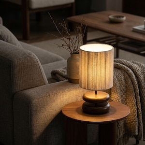 May include: A small, wooden table lamp with a cylindrical shade. The lamp sits on a round, wooden table next to a gray sofa. A vase with branches is visible in the background. The lamp's shade is a light beige color.
