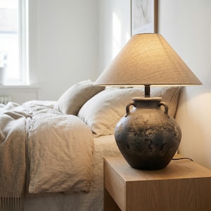 May include: A bedside lamp with a textured, dark gray ceramic base and a beige fabric lampshade. The lamp sits on a light wood nightstand next to a bed with beige bedding and pillows. The room has a neutral color palette.