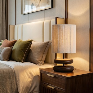 May include: A bedroom scene featuring a wooden nightstand with a lamp and a bed with pillows. The lamp has a beige shade and a dark brown base. The bed has several pillows in shades of brown and green.