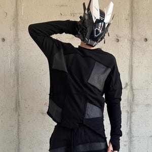 May include: Black long-sleeve top and pants with a patchwork design. The outfit includes a futuristic helmet with white and black accents. Gray high-top sneakers complete the look. The clothing has a distressed, deconstructed aesthetic.