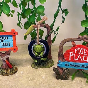May include: A whimsical fairy garden scene featuring three decorative signs. One sign reads "Pardon Our Pixie Dust", another has a frog in a tire swing, and the third says "Pixie Place, No Gnomes Allowed". The signs are brightly colored.