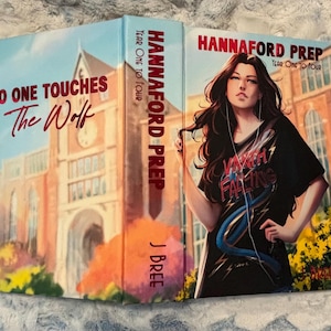 Op de afbeelding: Een hardcover boek getiteld Hannaford Prep Year One to Four door J Bree. De voorkant toont een illustratie van een persoon met lang donker haar in een zwart t-shirt. De achterkant toont een schoolgebouw met de tekst No One Touches The Wolf.
