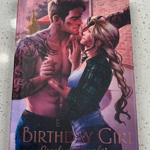 May include: A hardcover book titled Birthday Girl by Penelope Douglas featuring a romantic illustration of a shirtless man with arm tattoos and a woman with a long braid standing close together against a brick wall background in warm lighting.