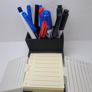Könnte beinhalten: Ein schwarzer Schreibtisch-Organizer mit Notizblock und Stifthalter. Der Stifthalter enthält blaue, rote und schwarze Stifte und Marker. Der Notizblock hat einen Stapel gelb liniertes Papier mit roten Linien und eine Reihe von Kreisen auf der linken Seite.