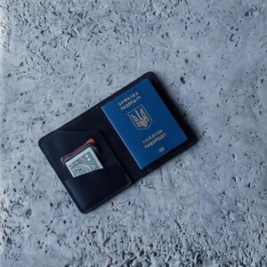 Puede incluir: Funda de pasaporte de cuero negro con un pasaporte ucraniano azul en el interior. El pasaporte tiene letras doradas y un emblema dorado. Unas cuantas tarjetas de crédito y un billete de banco son visibles en un bolsillo.