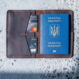 Puede incluir: Portapasaporte de cuero marrón abierto con un pasaporte ucraniano azul y dos tarjetas de crédito. El pasaporte tiene letras doradas y un emblema dorado. Las tarjetas de crédito están en un bolsillo en el lado izquierdo.