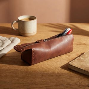 May include: A brown leather pencil case with a zipper, containing pens. A ceramic mug with coffee and a small book are also on the wooden surface. The pencil case is rectangular and has a stitched design.