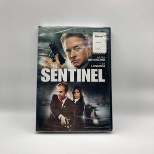 Puede incluir: DVD precintado de la película "The Sentinel". La portada presenta a Michael Douglas sosteniendo un arma, con Kiefer Sutherland y Eva Longoria también en la imagen. El título está en letras blancas grandes. Una pegatina de Walmart está en la esquina superior derecha.