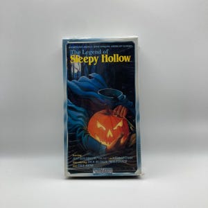 Puede incluir: Una cinta VHS de "La Leyenda de Sleepy Hollow" con una calabaza tallada y una figura encapuchada en la portada. La ilustración es azul y naranja, con el título en amarillo. La cinta está sellada.