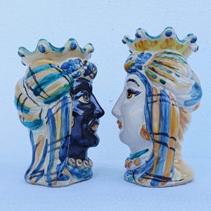 Può includere: Due sculture di teste in ceramica, una con volto blu scuro e l'altra bianca, entrambe ornate da corone e copricapi a turbante. Le sculture presentano dettagli dipinti in blu, giallo e bianco, mostrando una vista di profilo.