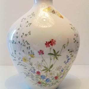 Könnte beinhalten: Eine weiße Porzellanvase mit engem Hals und goldfarbenem Rand. Der Körper der Vase ist mit einer zarten, farbenfrohen botanischen Illustration verschiedener Wildblumen, grüner Stängel und eines gelb-orangen Schmetterlings verziert.