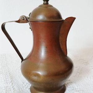 Può includere: Una brocca in metallo color rame con corpo arrotondato, base svasata, manico curvo e coperchio a cupola sormontato da un piccolo pomello. La superficie presenta una patina usurata con visibili variazioni di colore e consistenza nel metallo.