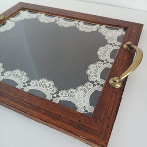 May include: A rectangular wooden serving tray with a dark gray interior and a decorative white lace border. The tray has brass handles on each side and a clear glass top.