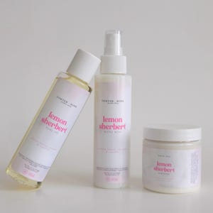 May include: Three white bottles of body care products with pink and white labels. The labels read "lemon sherbert" and "body oil", "milky mist", and "body lotion". The products are arranged on a white surface.