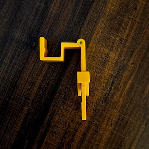 May include: A bright yellow, plastic, hinged object with a rectangular shape and a small, cylindrical hinge. The object is on a dark brown, wood-grain surface. The object appears to be a component of a larger item.