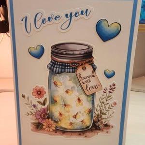 Könnte beinhalten: Eine Grußkarte mit hellblauem Rand, die ein Einmachglas voller leuchtender Glühwürmchen zeigt. Das Glas hat einen blau karierten Deckel, eine Schnur und ein Etikett mit der Aufschrift Preserved with love. Darüber steht der Text I love you, umgeben von blauen Herzen und bunten Blumen.