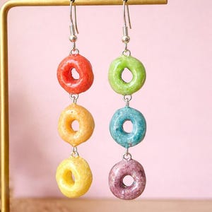 May include: Dangling earrings with colourful cereal-shaped charms. Each earring has three charms in red, orange, yellow, green, blue, and purple, connected by silver-toned metal. The earrings hang from silver-toned ear wires.